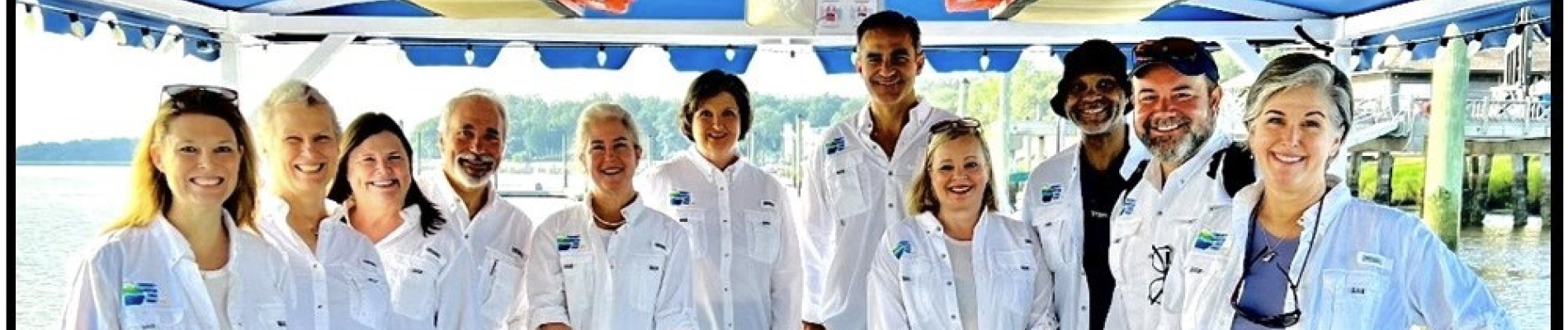Group of people on a boat, wearing white shirts, smiling under a blue-striped canopy.